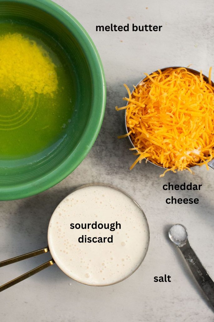 ingredients for sourdough cheddar cheese crackers on a table: melted butter, shredded cheese, sourdough discard, and salt.