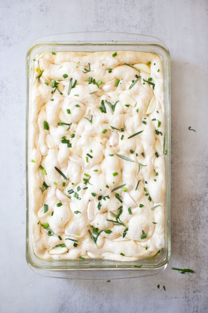 sourdough focaccia in a baking dish that has been dimpled and covered with herbs