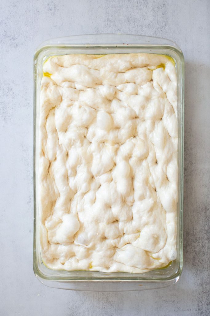 sourdough focaccia in a baking dish that has been dimpled and covered with olive oil 