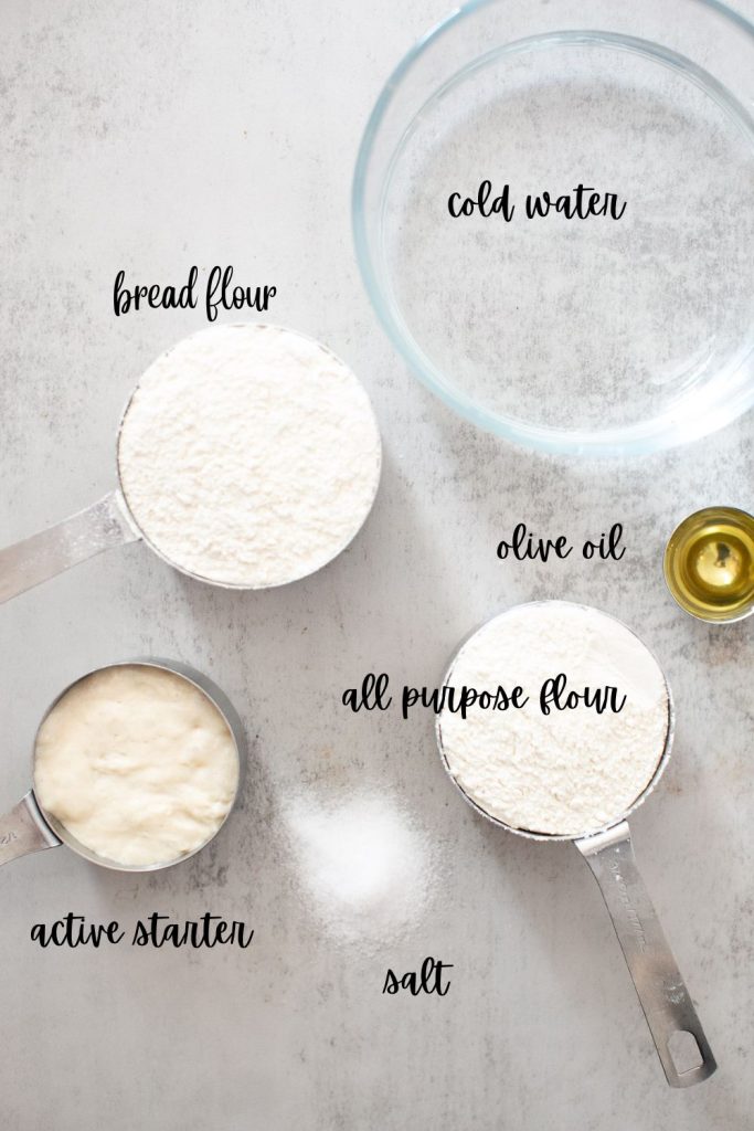 ingredients for sourdough dutch oven bread on a tray: active starter, bread flour, olive oil, salt, cold water, and all purpose flour
