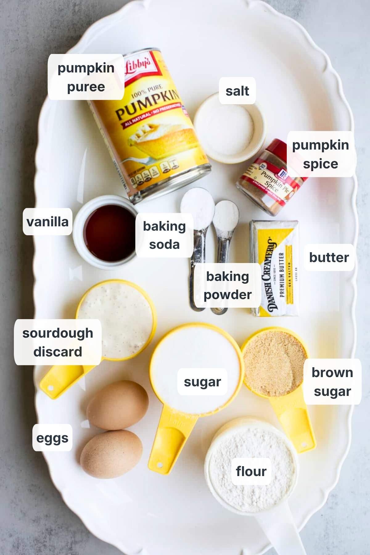 Ingredients for sourdough discard pumpkin streusel muffins on a tray: pumpkin puree, salt, vanilla, sourdough discard, pumpkin spice, butter, sugars, baking soda, and eggs.