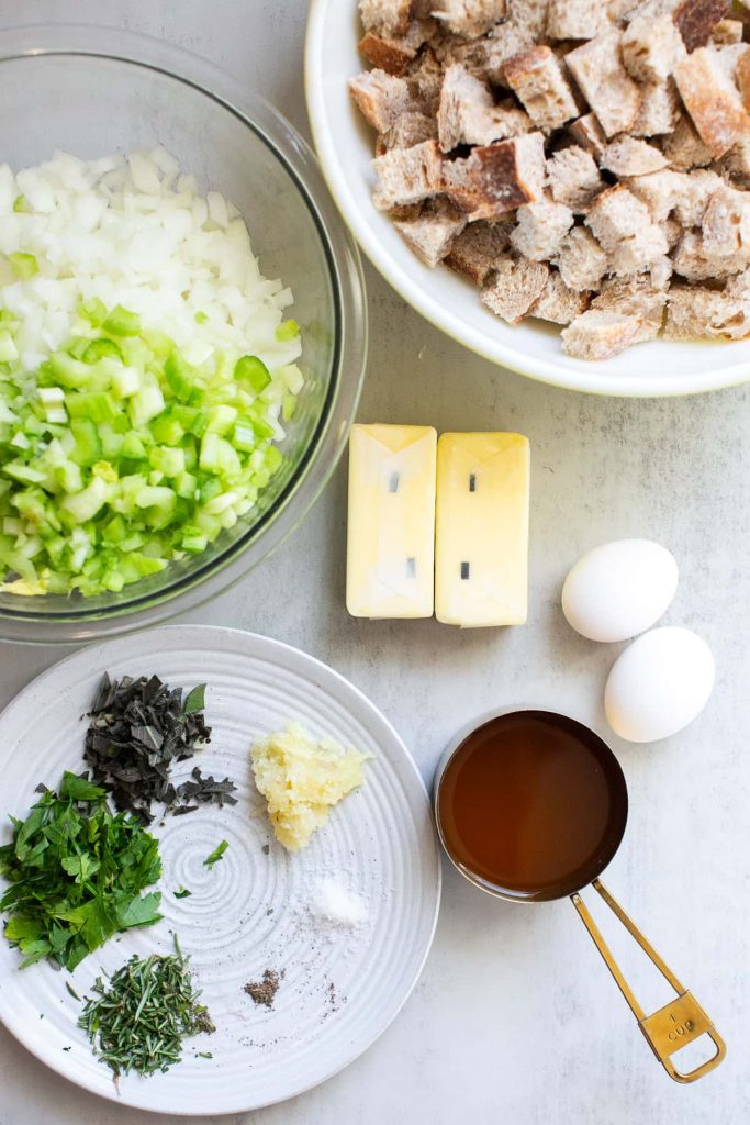 ingredients for sourdough stuffing on a plate: butter, celery, onions, eggs, herbs, garlic, and chopped sourdough bread