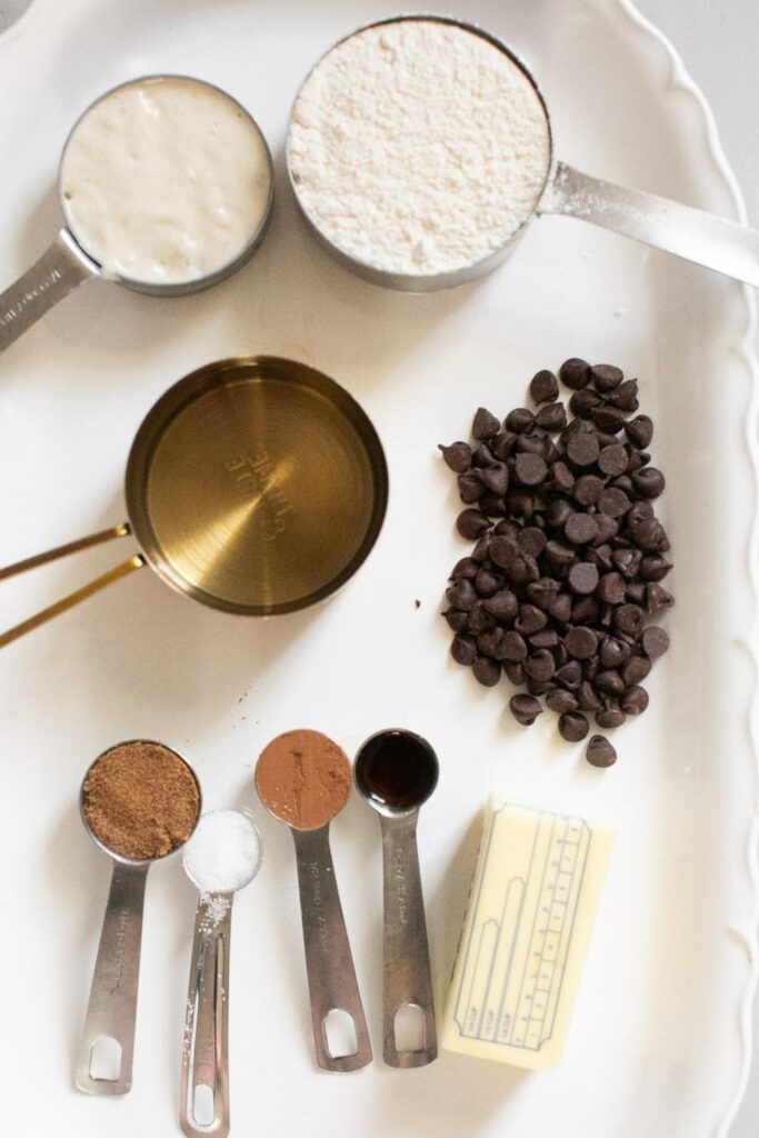 ingredients for sourdough chocolate focaccia bread on a plate: chocoalte chips, vanilla, cocoa powder, brown sugar, butter, water, sourdough starter, and bread flour