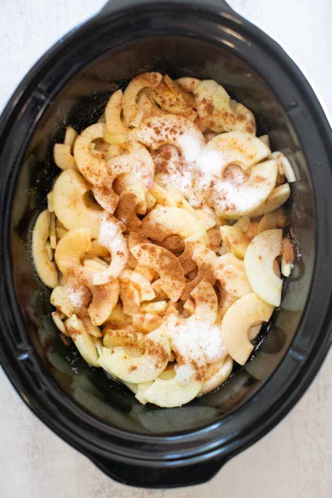 Chopped up apples in a crockpot on a counter with cinnamon and sugar on top.