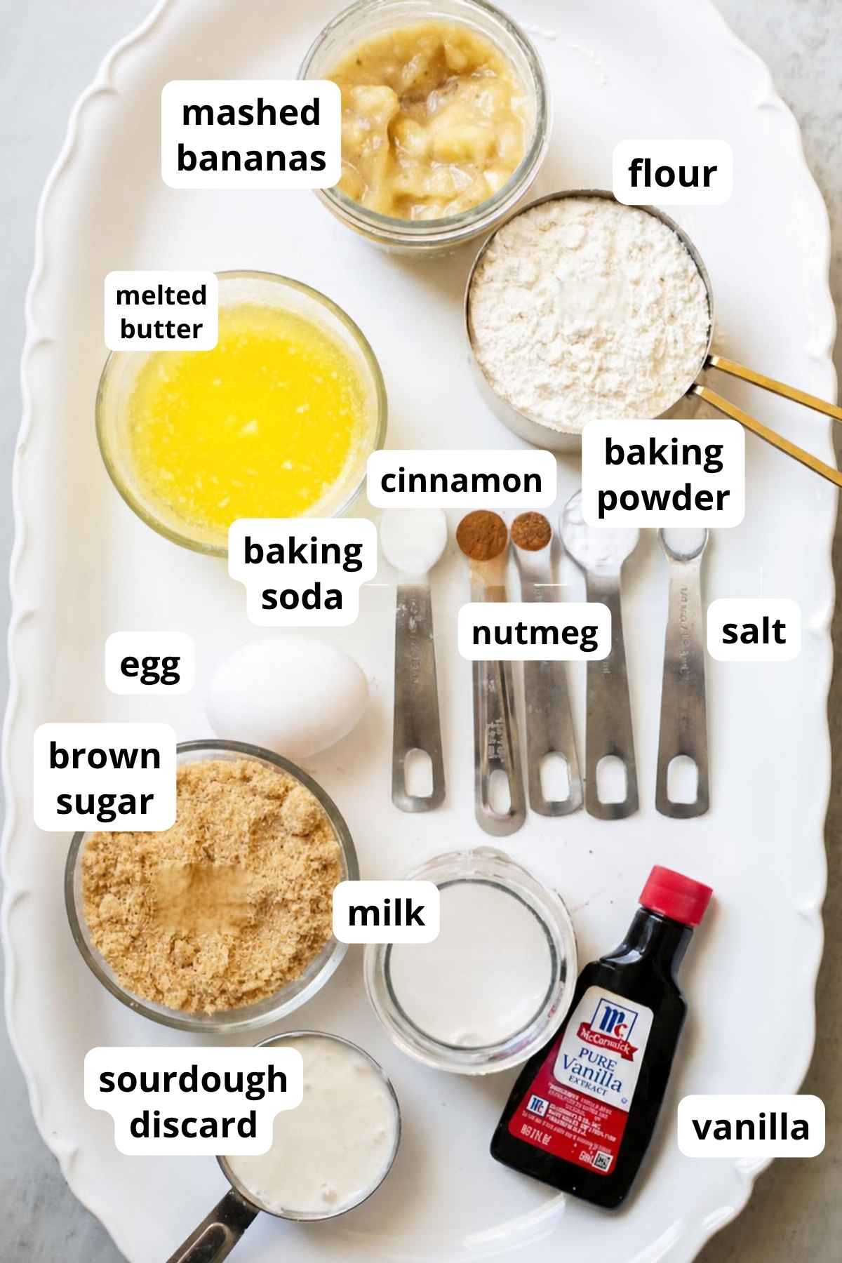 ingredients for sourdough discard banana muffins on a tray: egg, nutmeg, cinnamon, melted butter, mashed bananas, brown sugar, milk, vanilla, and sourdough discard