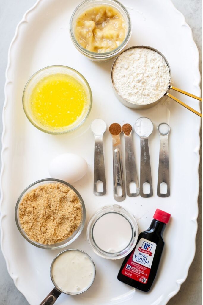 ingredients for sourdough discard banana muffins on a tray: egg, nutmeg, cinnamon, melted butter, mashed bananas, brown sugar, milk, vanilla, and sourdough discard